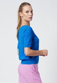 Suéter de punto azul de manga corta con patrón texturizado. Puños y dobladillo acanalados. Modelo combinado con pantalones rosas. Cuello redondo simple.