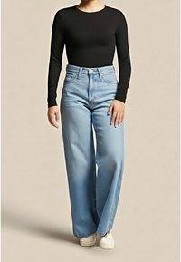 Personne portant un haut noir à manches longues, un jean large taille haute bleu clair et des chaussures blanches, debout devant un fond beige.