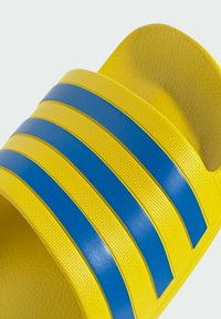 Zapatilla de piscina amarilla con tres franjas azules horizontales. Material texturizado con una superficie suave en la parte superior y un diseño de puntera abierta y redondeada.