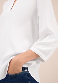 Personne portant une blouse blanche à manches longues avec un col en V, la main glissée dans la poche d'un jean bleu foncé, fond beige.
