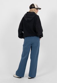 Femme portant un sweat à capuche noir, un pantalon large bleu, des chaussures blanches et une casquette de baseball beige et noire, debout devant un fond clair uni.