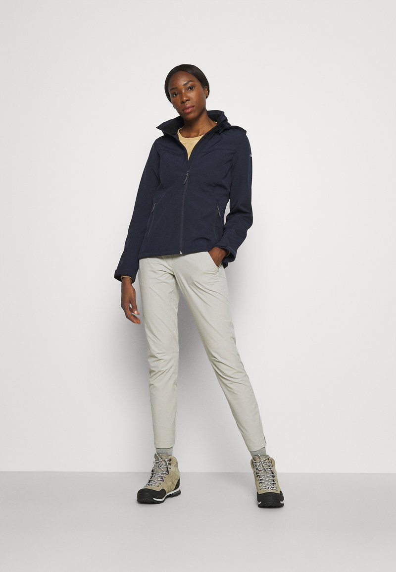 Veste imperméable bleu marine avec un col montant et une fermeture éclair sur le devant ; assortie à un pantalon beige clair et des chaussures de randonnée grises avec des accents noirs.