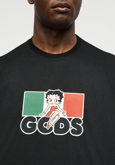 Μαύρο πουκάμισο με μια καρτούν γυναίκα σε κόκκινο φόρεμα που κάθεται πάνω στη λέξη «GODS» μπροστά σε ένα τρικολόρ φόντο πράσινου, λευκού και κόκκινου χρώματος.