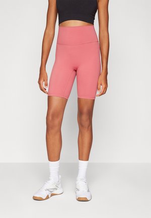 Vysoké, růžové cyklistické šortky vyrobené z pružného materiálu s plochými švy, doplněné o černý crop top a bílé sportovní boty.