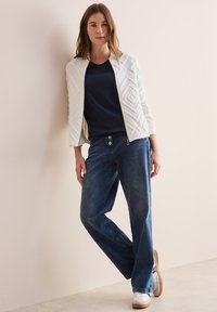 Witte gestikte jas met geometrisch patroon, marineblauw T-shirt, blauwe jeans en witte sneakers. Model staat tegen een neutrale achtergrond.