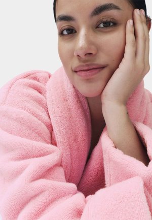 Joven mujer con un suave albornoz rosa, apoyando su rostro en la mano, con una expresión tranquila y una ligera sonrisa sobre un fondo blanco.