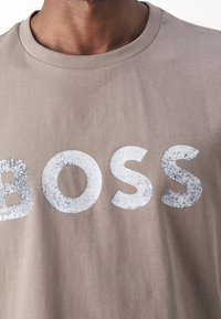 Detailní záběr na béžové tričko s opotřebovanými bílými písmeny, která na hrudi vypisují "BOSS", na osobě s tmavou pletí.