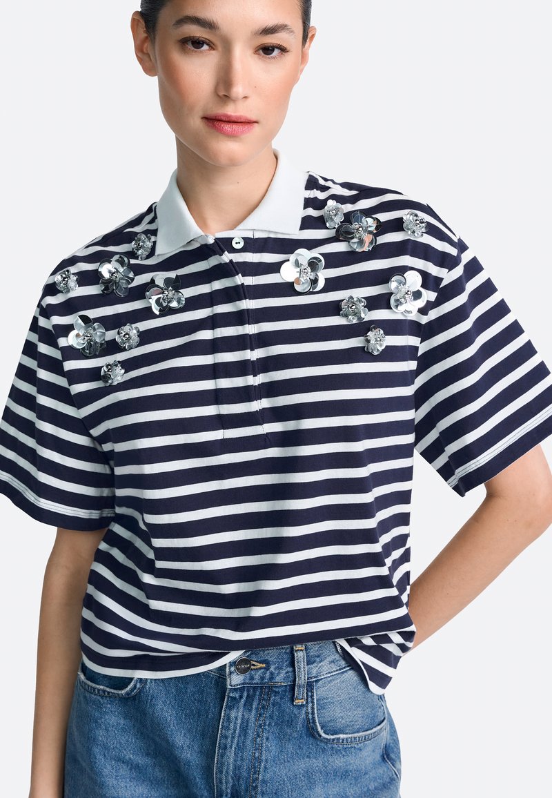 Polo de rayas azul marino y blanco con mangas cortas, con adornos florales de lentejuelas en la parte delantera y un cuello clásico.