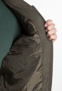 Main tenant une veste verte ouverte montrant la poche intérieure zippée et la doublure en tissu lisse.