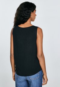 Mujer con cabello rizado, vestida con una blusa negra sin mangas y jeans azules, vista desde atrás contra un fondo claro y sencillo.