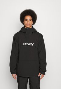 Czarna wodoodporna kurtka z kapturem, z dużą kieszenią z przodu i przerośniętym białym logo "OAKLEY". Teksturowany, lekki materiał.