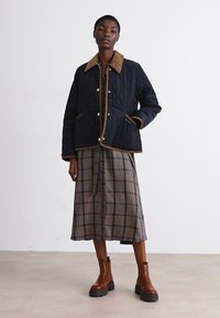 Casaco acolchoado azul-marinho com gola castanha, apresentando botões de pressão e bolsos aplicar, usado sobre uma saia xadrez e botas de tornozelo castanhas.