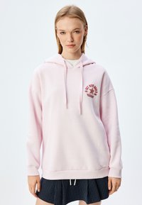Sudadera de gran tamaño en rosa claro, fabricada con un tejido suave, que cuenta con capucha ajustable y un gráfico circular con una estrella roja y texto.