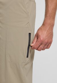 Pantaloni beige leggeri con una finitura testurizzata, dotati di una tasca con zip nera e di una tasca laterale a toppa. Primo piano di una mano che apre la zip.