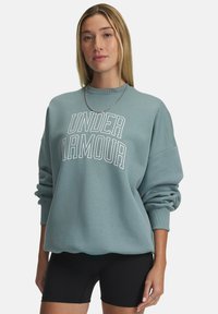 Lichtblauwe sweatshirt met "UNDER ARMOUR" in het wit gedrukt, met een ronde hals en ingezakte schoudernaden, gecombineerd met zwarte shorts.