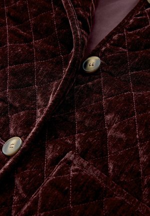 Burgundy quilted velvet fabric with a soft texture, featuring a patch pocket and round, metallic buttons on the front.