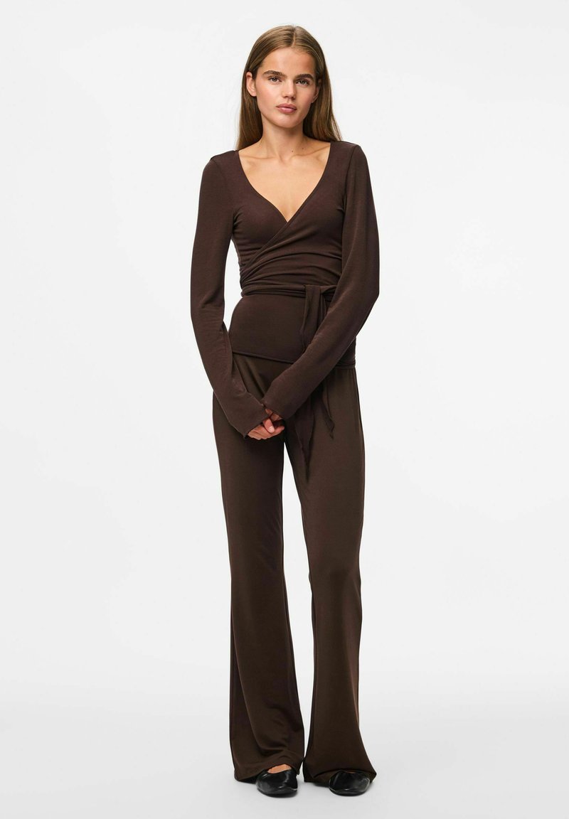 Brūns garām piedurknēm apģērba topiņš ar V veida kaklu un sasietu jostasvietu, apvienots ar atbilstošām platām biksēm. Mīksts, elastīgs audums ar gludu tekstūru.