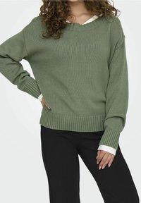 Zielony dzianinowy sweter z dekoltem w serek, ściągaczami przy rękawach i u dołu. Nałożony na białą koszulę. W połączeniu z luźnymi czarnymi spodniami.