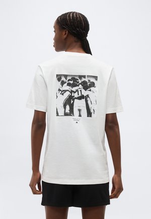 Woman with braided hair wearing a white T-shirt showing a black-and-white photo of a group huddle with arms around each other on the back.