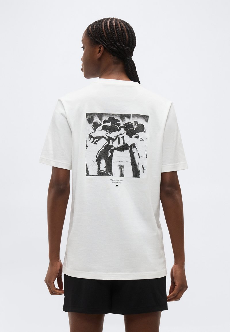 Γυναίκα με πλεξούδες που φοράει λευκό μπλουζάκι και δείχνει μια ασπρόμαυρη φωτογραφία μιας ομαδικής αγκαλιάς με τα χέρια γύρω από τους άλλους στην πλάτη.