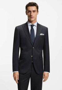 Granatowy garnitur w prążki z zapięciem na jeden guzik, klapami z wycięciem i przednimi kieszeniami. Biała koszula z kołnierzykiem oraz krawat w groszki. Szczegół w postaci poszetki.