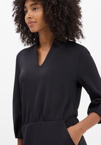 Schwarzes V-Ausschnitt-Kleid mit langen Ärmeln, gefaltetem Taillengürtel und seitlichen Taschen, aus glattem, leichtem Material, mit einem minimalistischen Design.