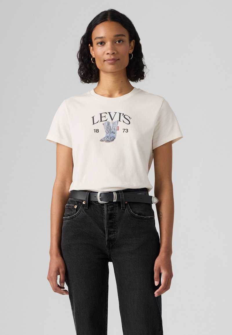 Γυναίκα με σγουρά μαλλιά φοράει λευκό t-shirt Levi's με γραφικό μποτάκι, μαύρα τζιν και μαύρη ζώνη, στέκεται μπροστά σε απλό γκρι φόντο.