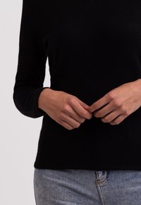 Sort langærmet top med blød tekstur og rund halsudskæring, parret med lyseblå slidte denimjeans. Hænderne placeret på hoften.