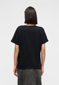 Γυναίκα με σκούρα μαλλιά μήκους έως τους ώμους, που φοράει μια απλή μαύρη κοντομάνικη μπλούζα και ξεθωριασμένη μαύρη φούστα από τζιν, στέκεται με την πλάτη προς τα εμπρός.