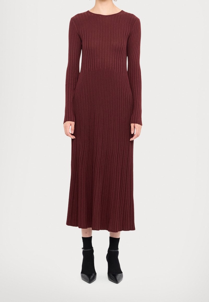 Femme portant une robe midi bordeaux côtelée à manches longues associée à des bottines noires sur un fond blanc uni.