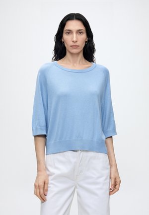 Femme aux longs cheveux foncés portant un haut bleu clair à manches courtes et un pantalon blanc, debout devant un fond blanc uni.