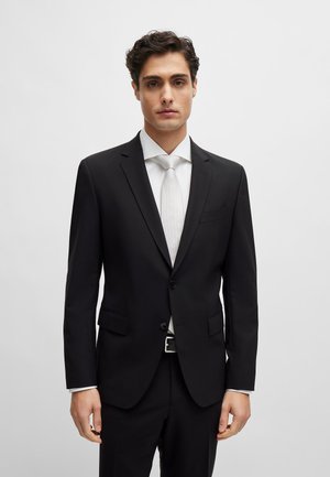 Chaqueta de traje negra con corte slim, textura suave, solapas de muesca y cierre de dos botones, combinada con una corbata de color claro y una camisa blanca.