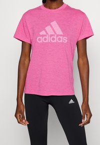 Camiseta rosa de manga corta hecha de una tela texturizada con un gran logo blanco de Adidas en el pecho. Combinada con leggings negros.