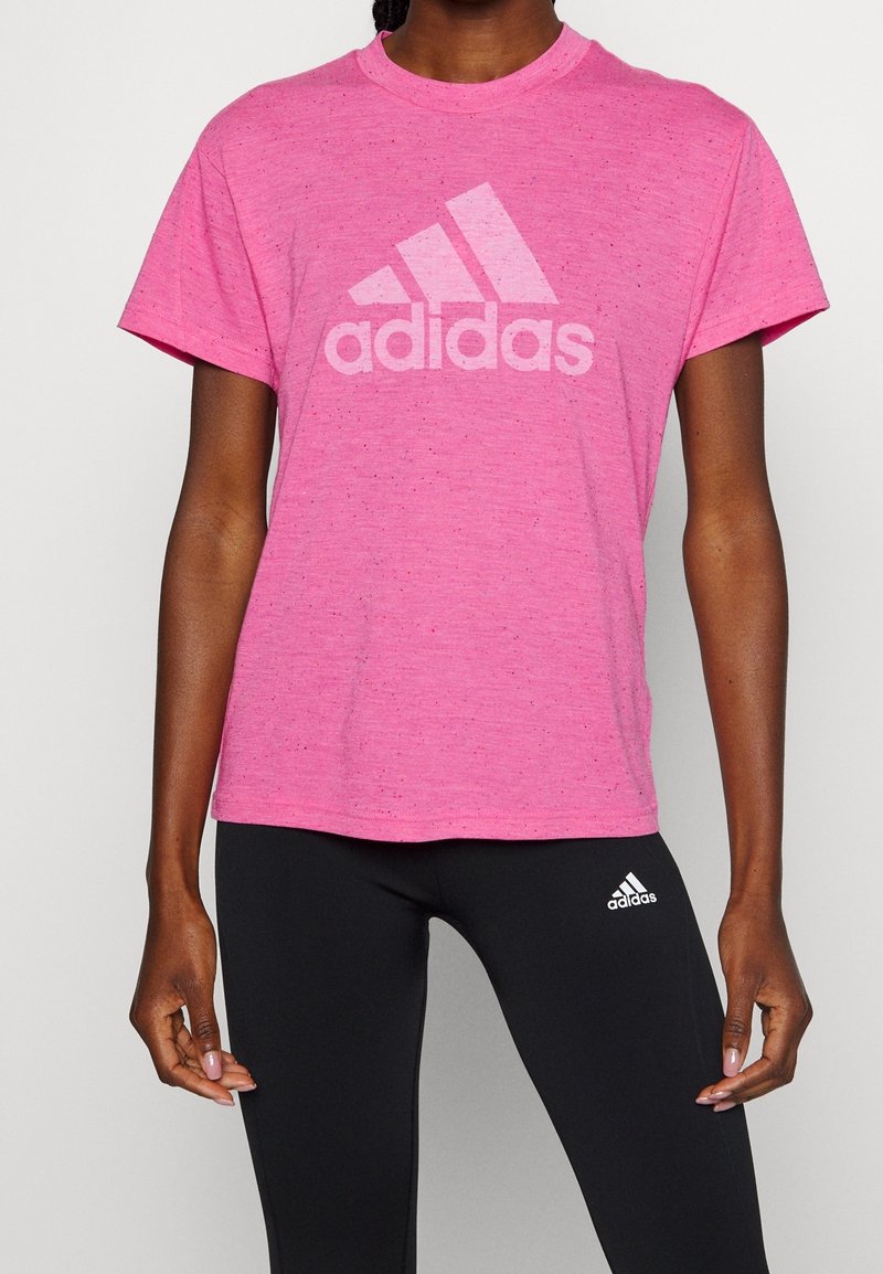 Camiseta rosa de manga corta hecha de una tela texturizada con un gran logo blanco de Adidas en el pecho. Combinada con leggings negros.