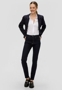 Marineblazer, wit overhemd met knopen, donkere jeans en zwarte instappers met gouden kettingdetail. De blazer heeft een gestructureerd ontwerp en een gestroomlijnde pasvorm.