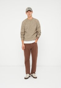 Maglione in maglia beige con motivo testurizzato, T-shirt bianca sottostante, pantaloni marroni, berretto grigio e sneakers bianche con dettagli.
