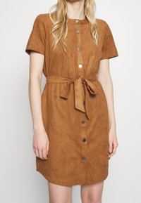 Robe chemise en faux suede marron avec des manches courtes, un devant à boutons et une ceinture à nouer à la taille. Texture douce avec une coupe décontractée.
