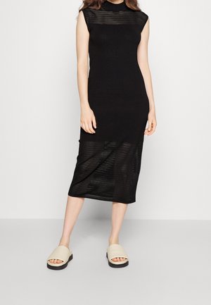 Femme portant une robe midi noire sans manches avec des panneaux rayés transparents et des sandales beiges à enfiler, debout devant un fond clair uni.