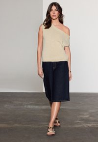 Beige één-schouder gebreide top, donkere denim culottes en gemusterde sandalen, met lang donker haar en oorbellen, staand op een grijze vloer.