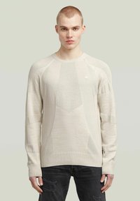 Beige gestrickter Pullover mit strukturierten Einsätzen, gerippten Bündchen und Rundhalsausschnitt. Über dunklen Jeans getragen. Leicht gemusterter Stoff.