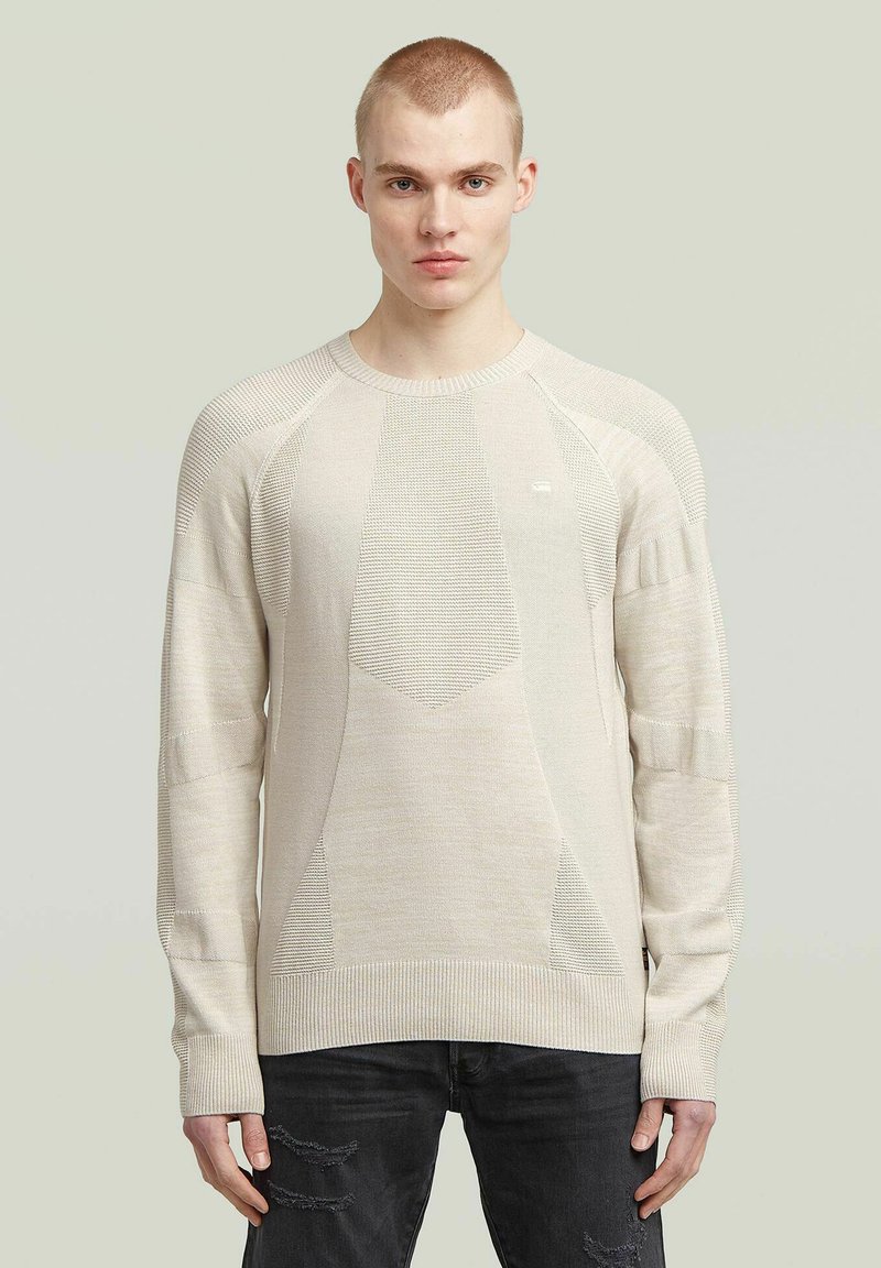 Beige gestrickter Pullover mit strukturierten Einsätzen, gerippten Bündchen und Rundhalsausschnitt. Über dunklen Jeans getragen. Leicht gemusterter Stoff.