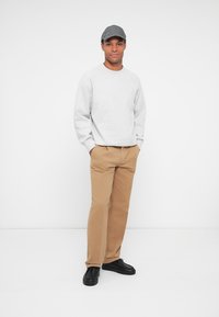 Sweatshirt cinza, calças bege e sapatos pretos; modelo usa um boné cinza; postura em pé, fundo neutro.