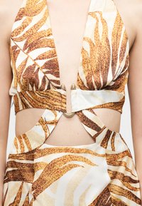 Robe à col halter avec un décolleté en V profond, une boucle dorée à la taille, et un motif de feuilles tropicales orange-brun sur tissu blanc, avec des découpes au niveau du ventre.