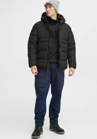 Geacă de puf neagră cu glugă, strat negru dedesubt, pantaloni cargo bleumarin și boots negre; căciulă tricotată gri; buzunare funcționale pe pantaloni.