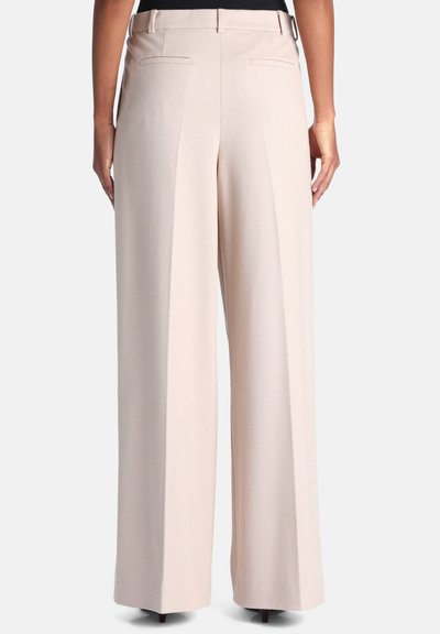 Femme portant un pantalon beige à jambes larges, debout, les mains le long du corps, vue de dos sur un fond uni.
