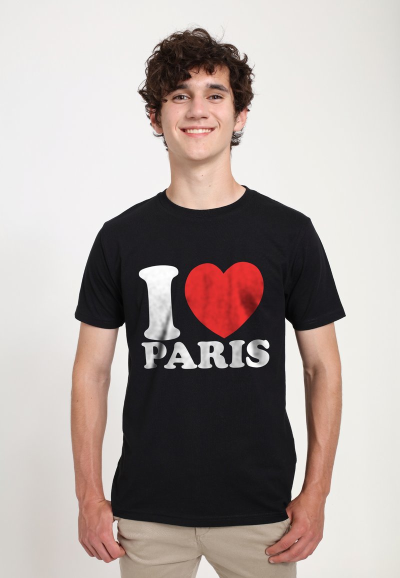 Mladý muž s kudrnatými vlasy se usmívá, má na sobě černé tričko s bílým a červeným nápisem "Milujem Paříž", stojí proti jednoduchému světlému pozadí.