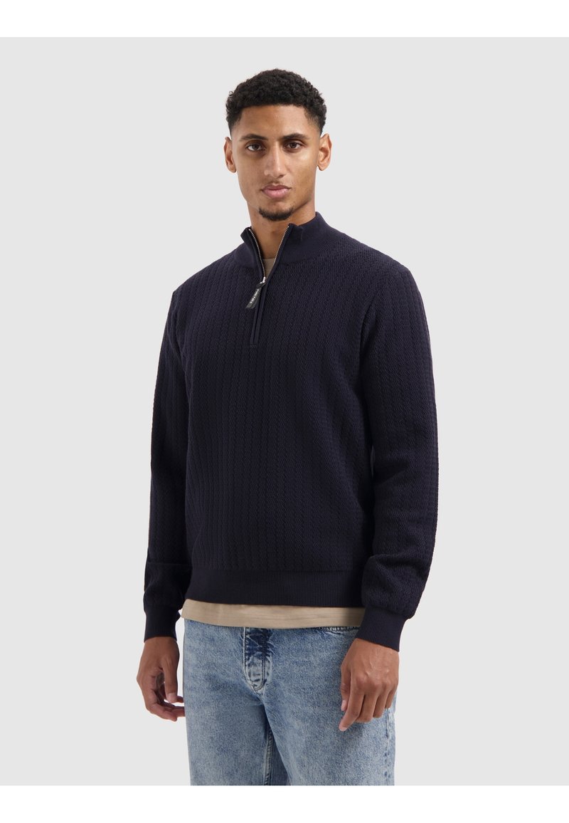 Marineblauw ritsvest met een textuurgebreide patroon, geribde manchetten en zoom, gedragen over een beige shirt, in combinatie met lichtblauwe jeans.
