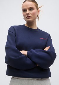 Tumesinine pusa ümmarguse kaelusega, langetatud õlgedega ja soonikkoes varrukaservadega. Rinnal väike punane logo. Pehme tekstuur.