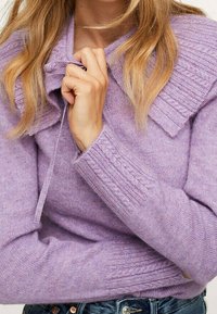 Donna che indossa un maglione lilla lavorato a maglia con colletto arricciato, tenendo il laccetto del colletto vicino al collo, abbinato a jeans blu.