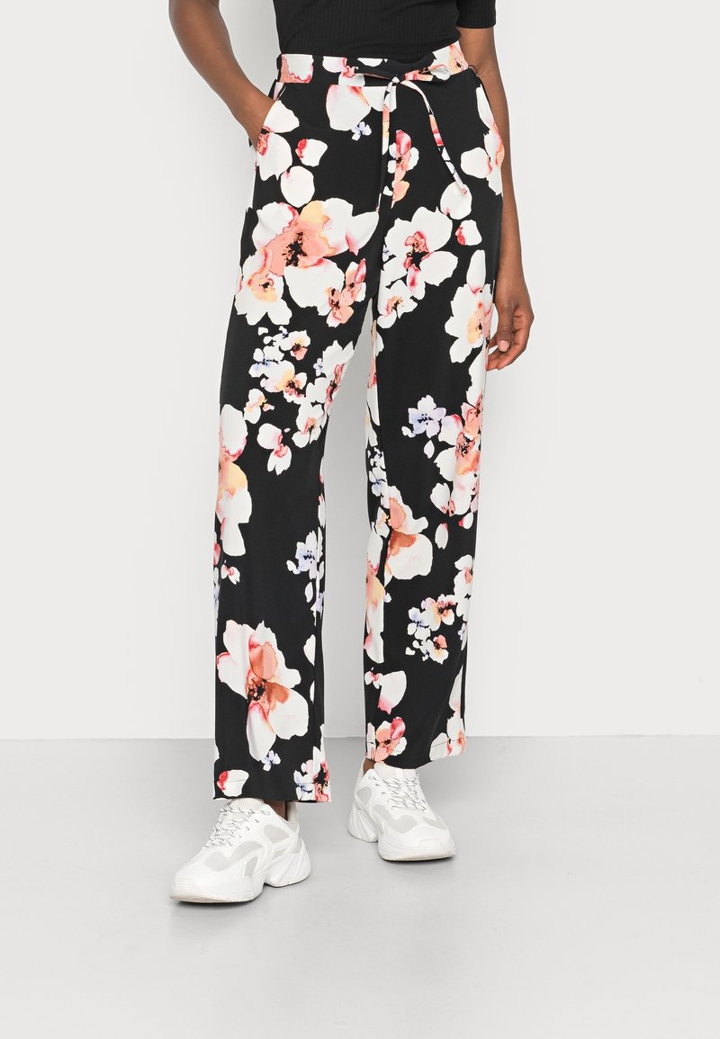 Personne portant un pantalon noir à jambes larges avec de grands motifs floraux roses et blancs, un haut noir et des baskets blanches épaisses, debout les mains dans les poches.